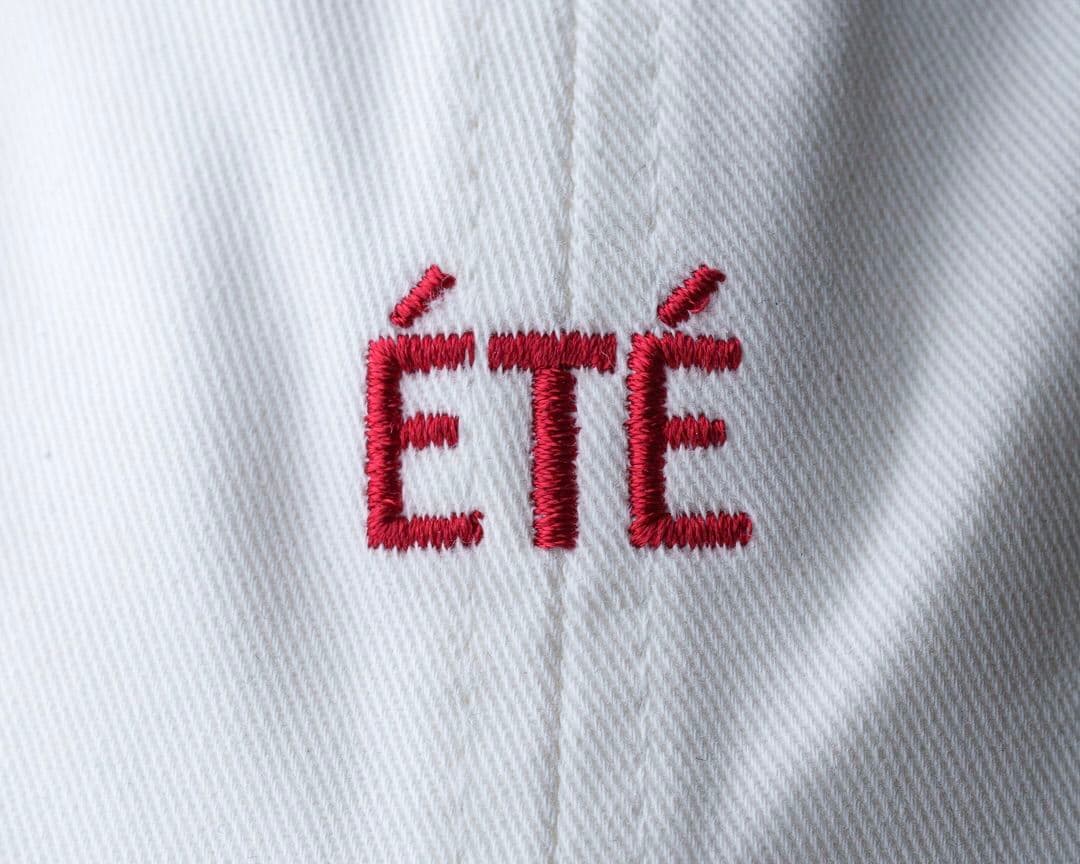 Close-up of the word "ÉTÉ" embroidered in red on white fabric with a textured pattern.
