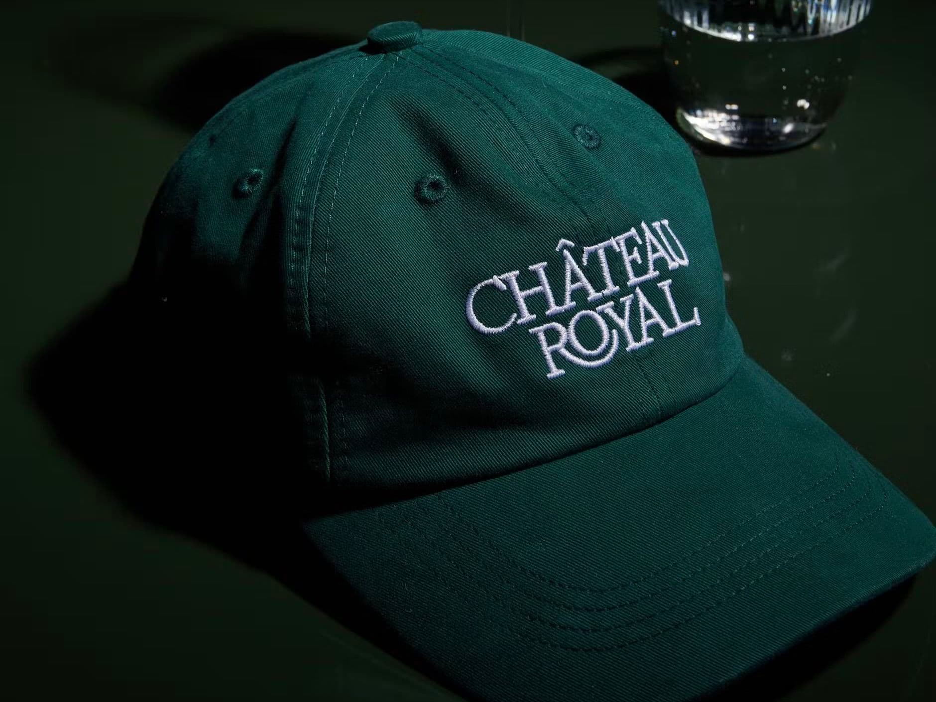 Château Royal green custom embroidered cap on a dark table beside a wine glass and water glass.