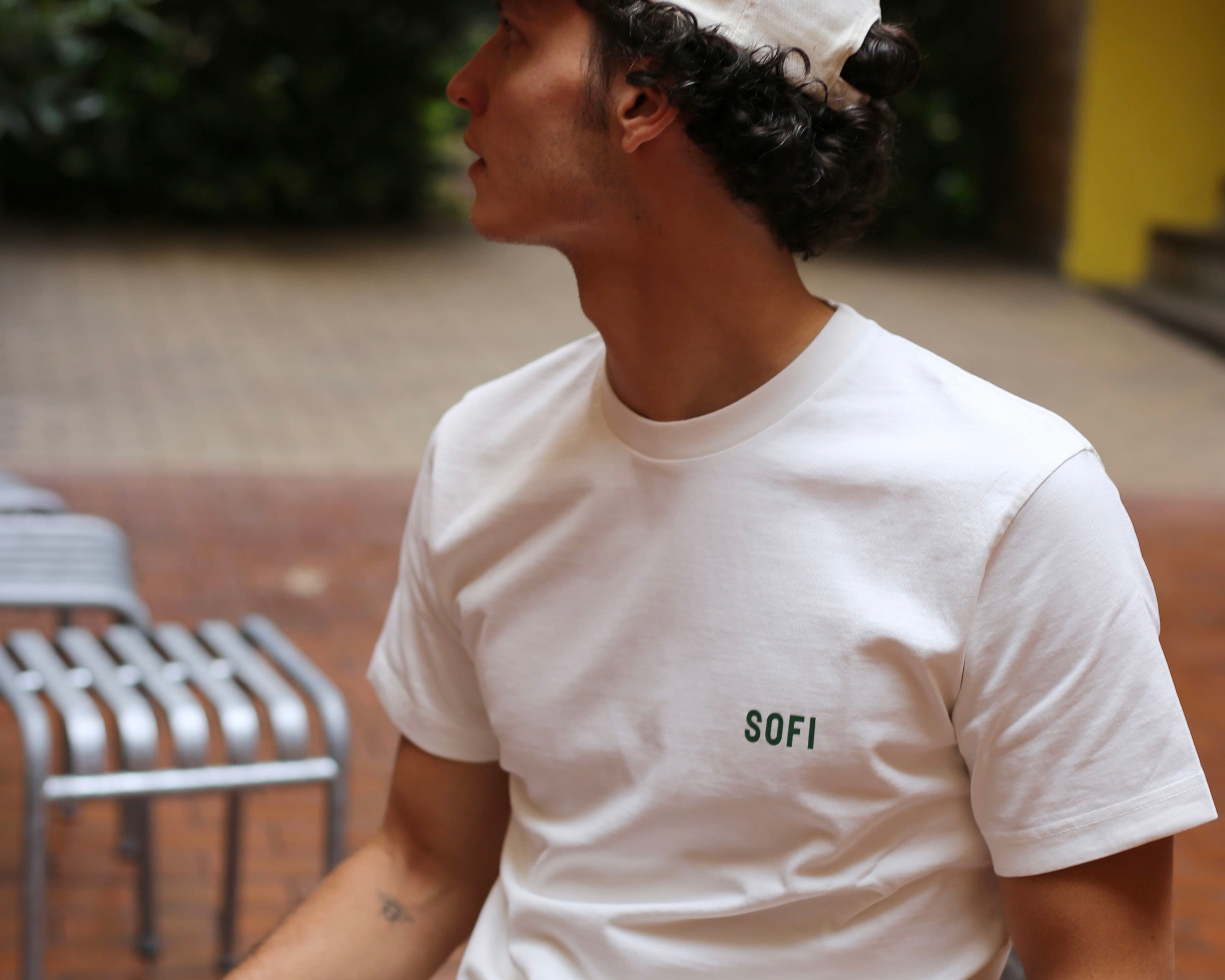 A person wearing a white T-shirt with "SOFI" text, sitting outdoors on a bench, looking to the side.
