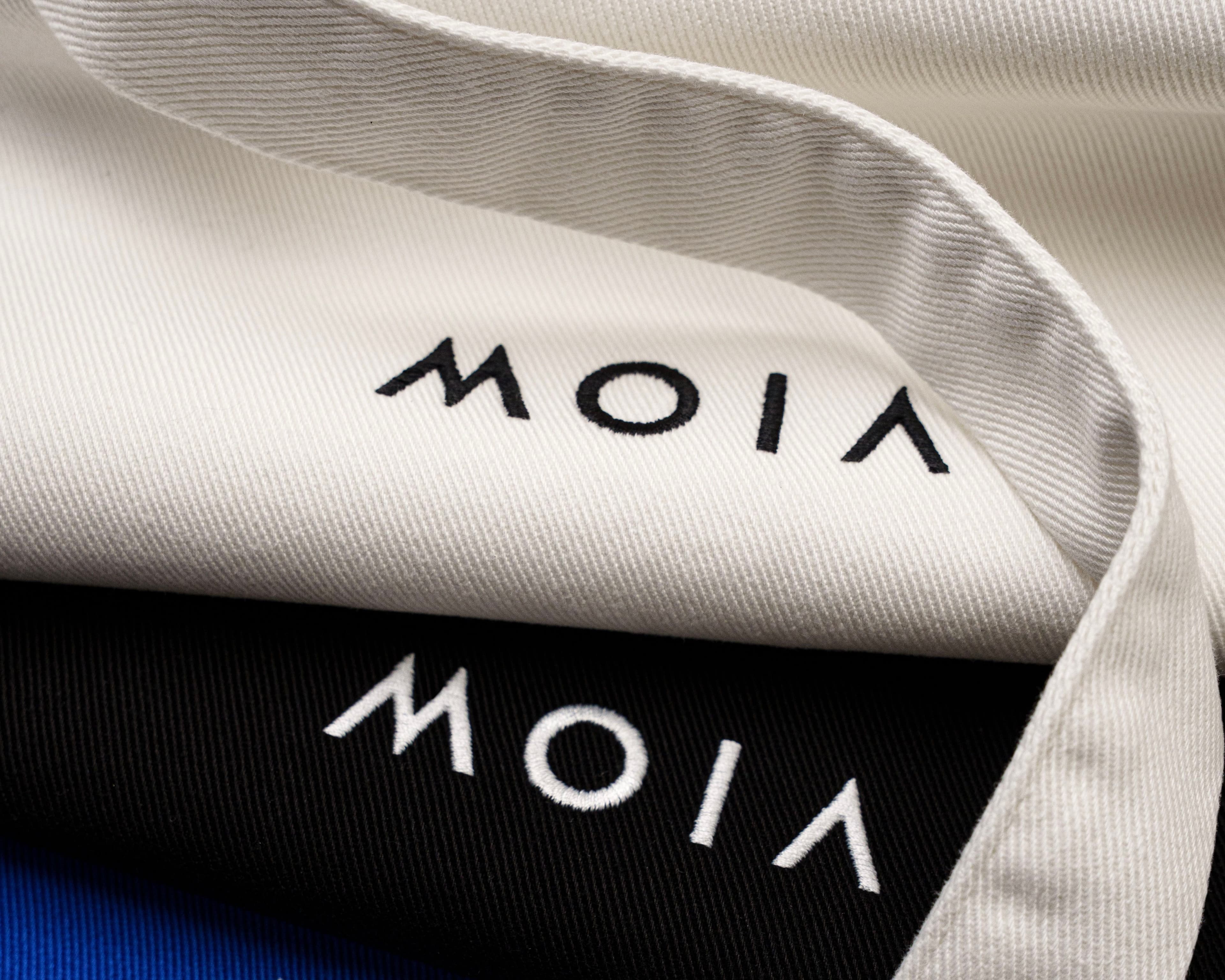 Close-up of embroidered logos "MOIA" on cotton tote bags in white, black, and blue fabric.