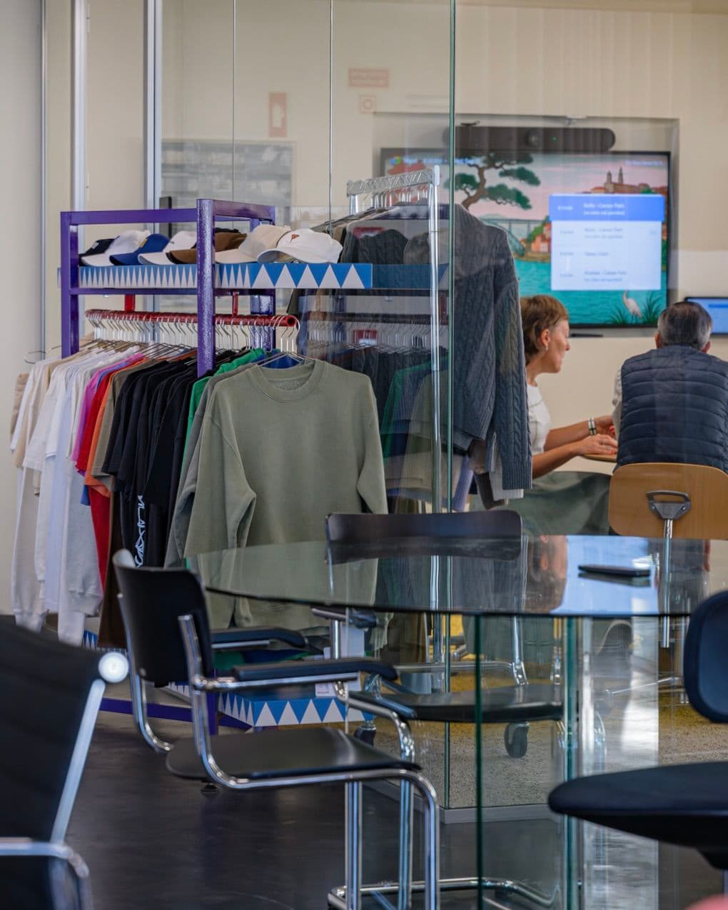 Glass-walled office with a clothing rack displaying shirts and sweaters. Two people are seated at a table with a screen in the background.
