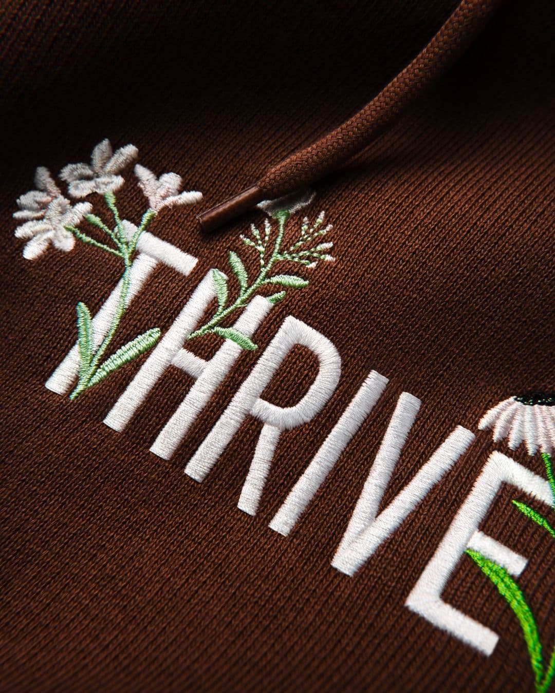 Close-up of embroidery on a brown hoodie with the word Thrive stitched in white and floral details in green and white thread.