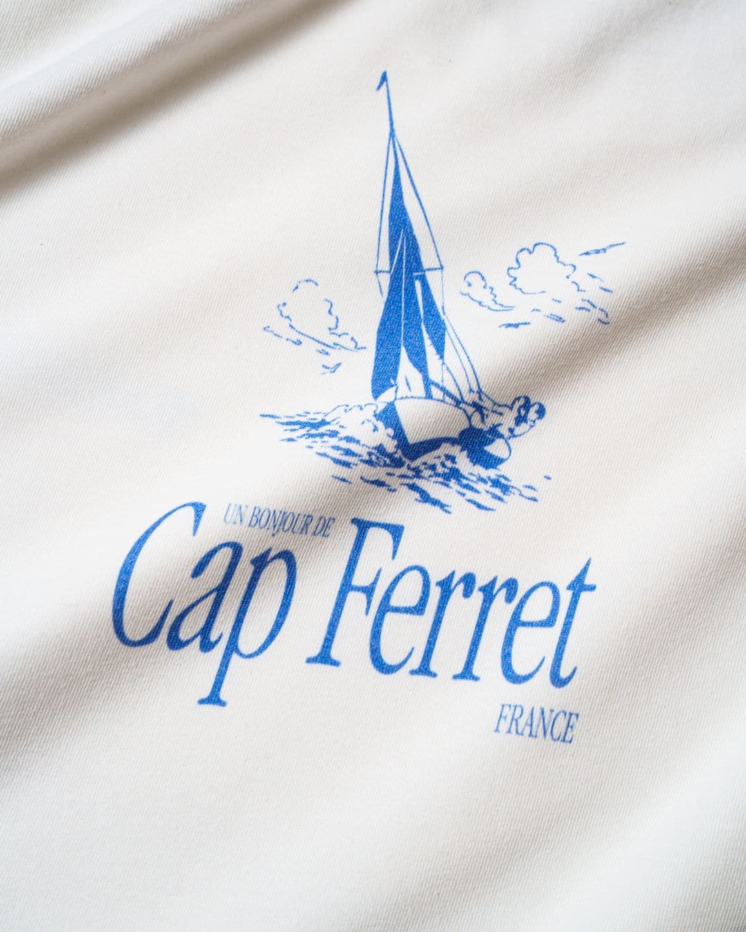 White fabric with a blue sailboat illustration and text reading "Un Bonjour de Cap Ferret France."