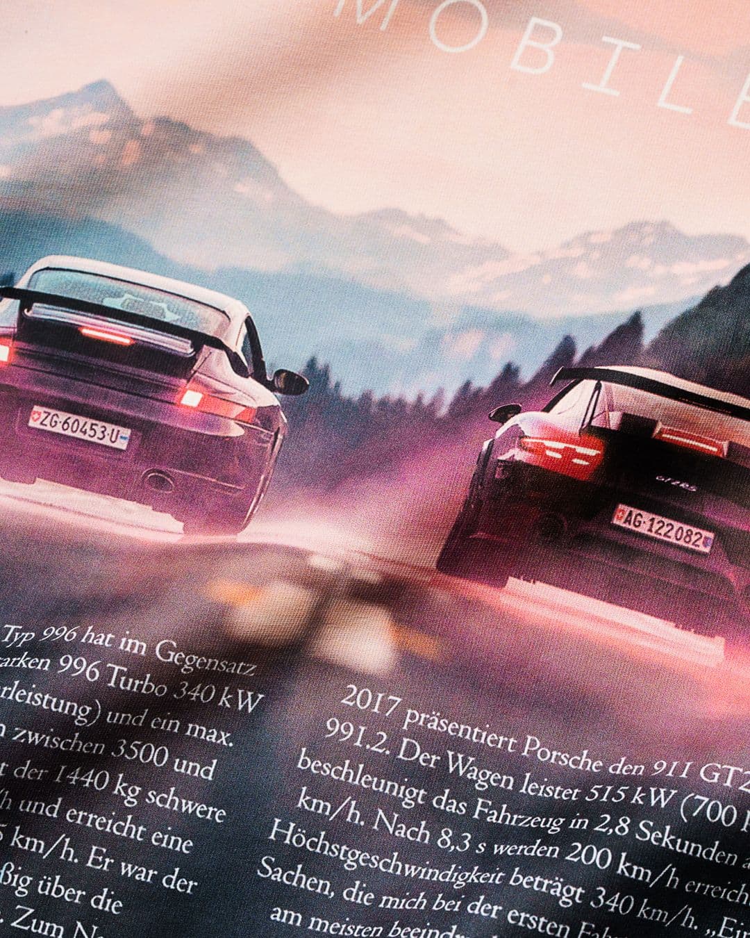 Two sports cars driving on a road with mountains in the background. Text in German is visible at the bottom of the image.