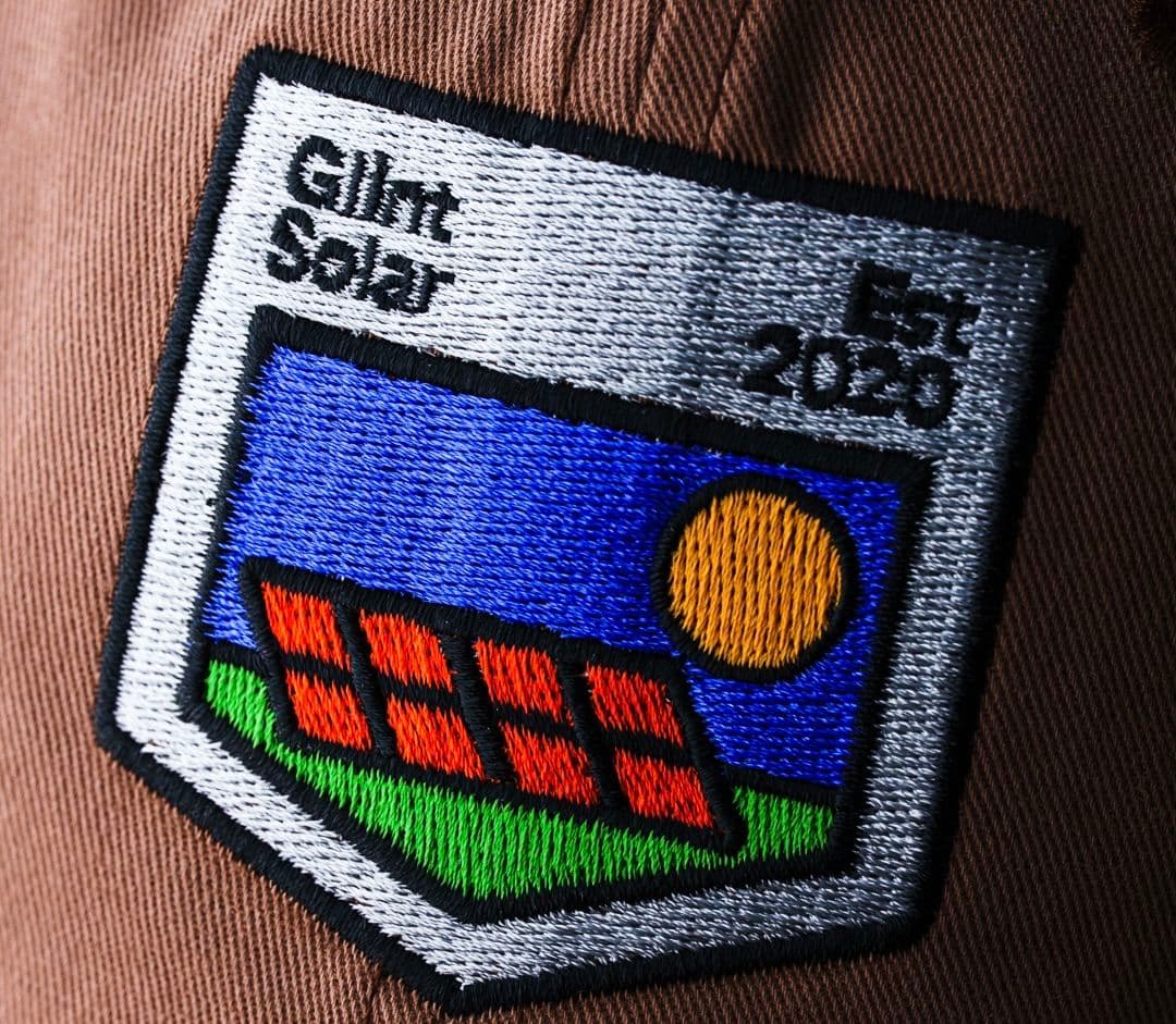 Embroidered patch with "Glint Solar Est 2020," featuring solar panels, green field, and a sun on a brown fabric background.