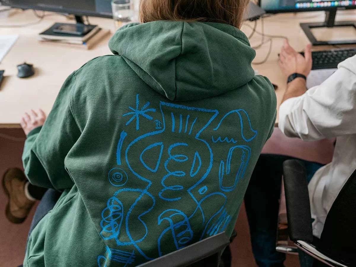Person wearing a green In August custom hoodie with blue graphic back print, seated at a desk in an office setting