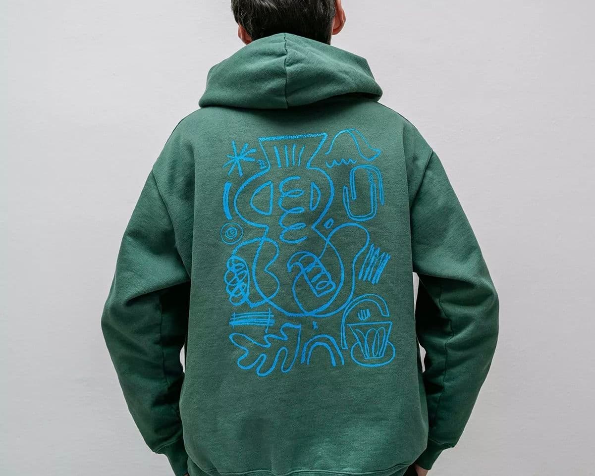 In August employee wearing a custom green hoodie with abstract blue line art on the back, standing against a plain white background.