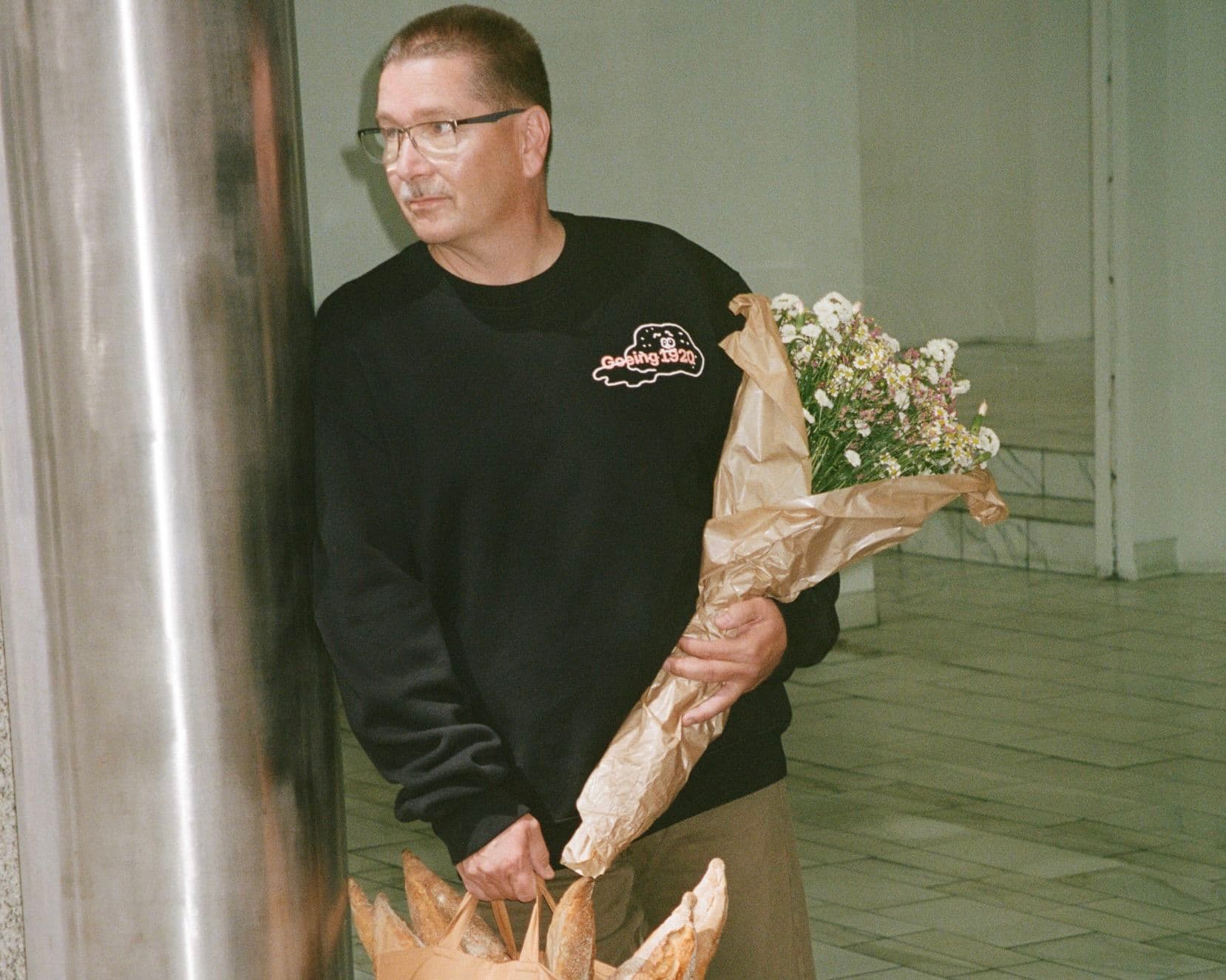 Man holding a bouquet of flowers and a paper bag with baguettes, standing near a metal column in a tiled indoor space wearing a black sweatshirt with the word Goeing 1920.