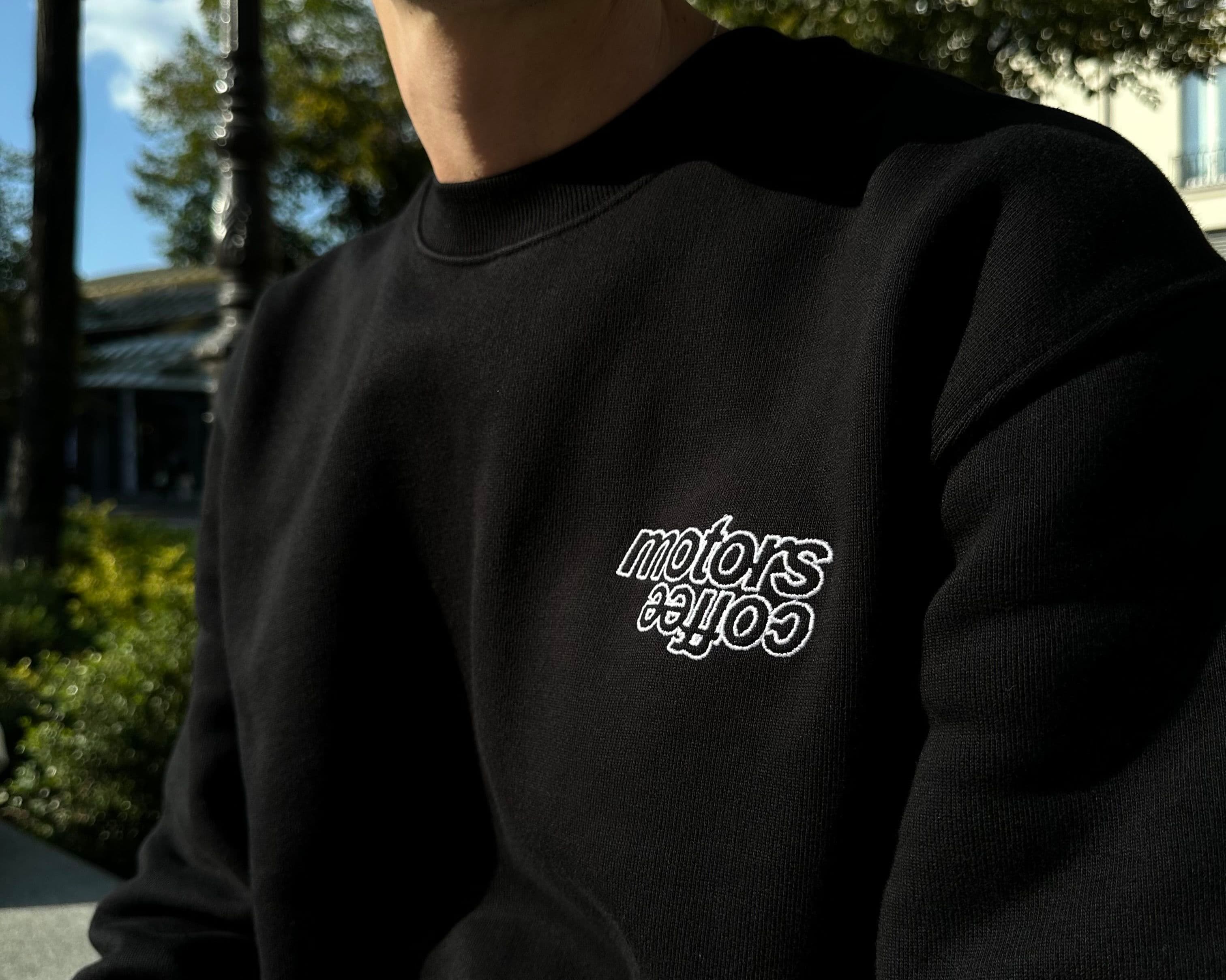 Person wearing a black sweatshirt with "coffee motors" embroidered in white, sitting outdoors in sunlight with trees in the background.