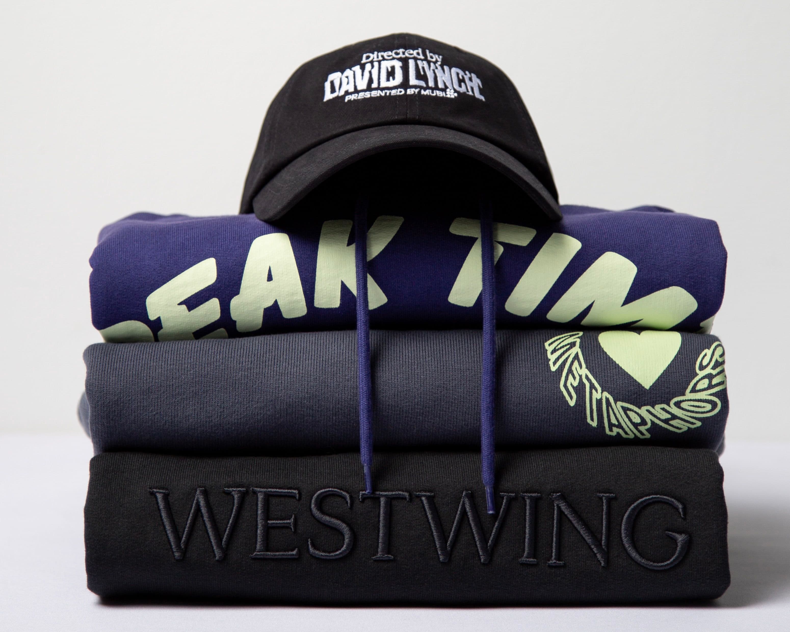 A stack of folded sweatshirts with "PEAK TIME" and "WESTWING" text, topped with a black cap reading "Directed by DAVID LYNCH."