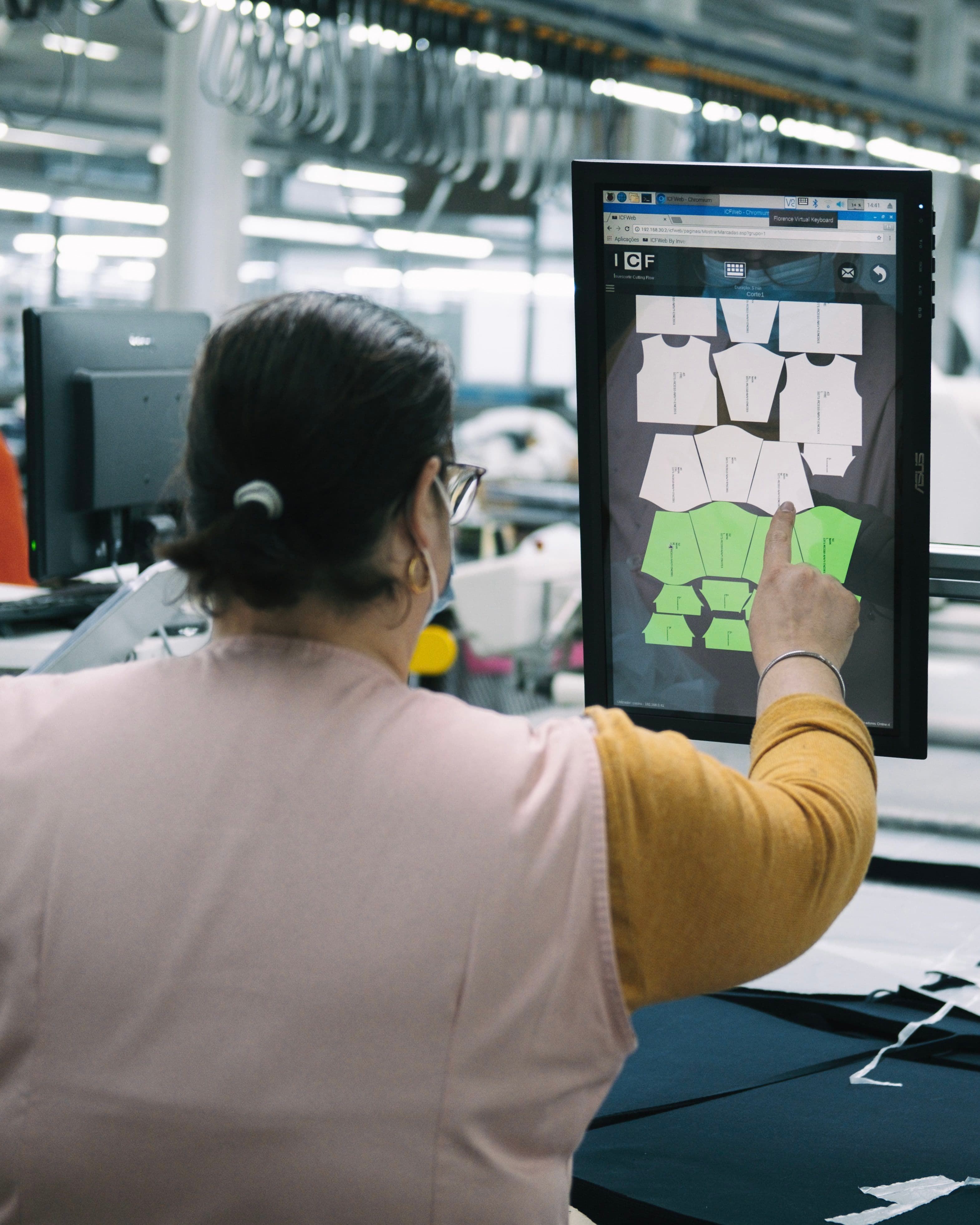 Person using a touchscreen to select clothing patterns in a garment factory, with fabric pieces displayed on the screen.