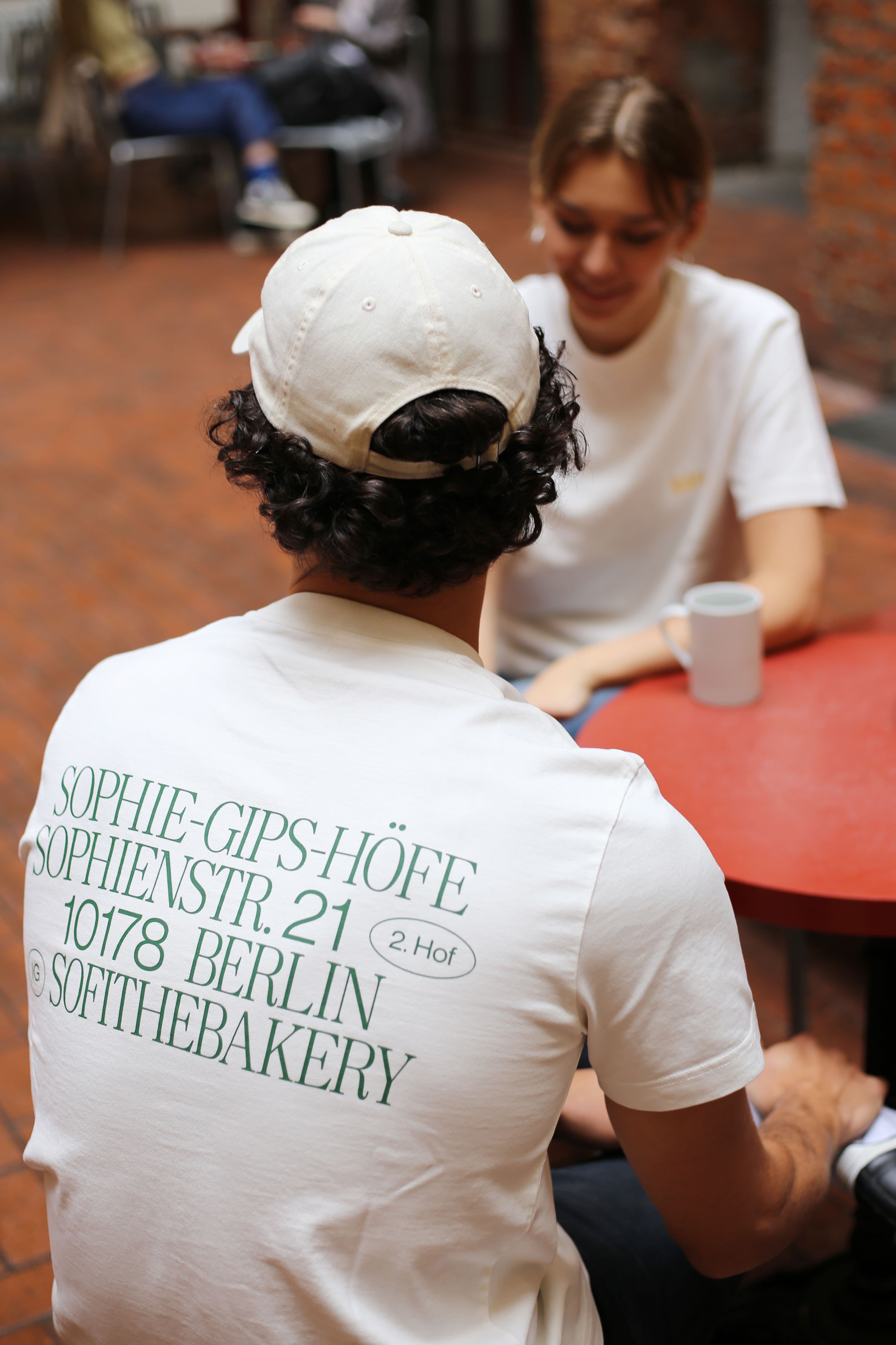 Two people wearing branded t-shirts and cap, sitting at a café table with a mug on the table.