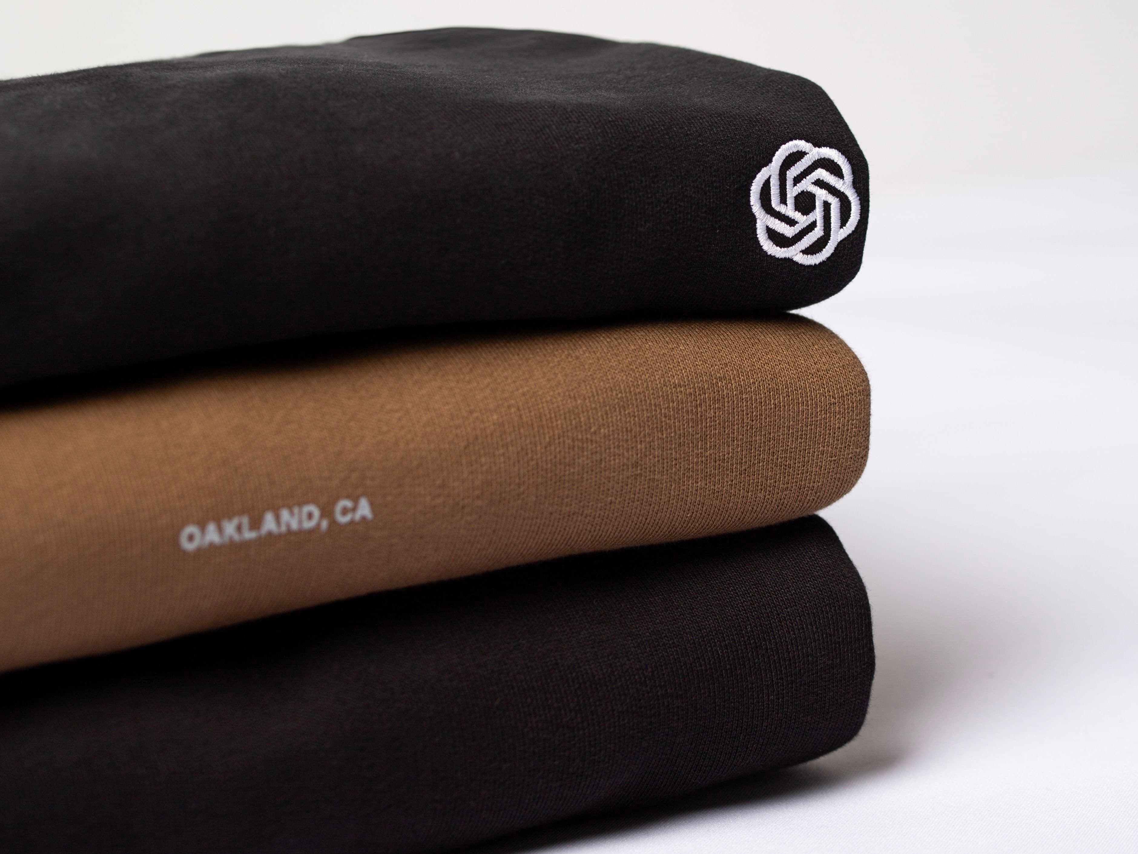 Stack of folded shirts: top and bottom black with a white logo, middle brown with "OAKLAND, CA" text, on a white surface.