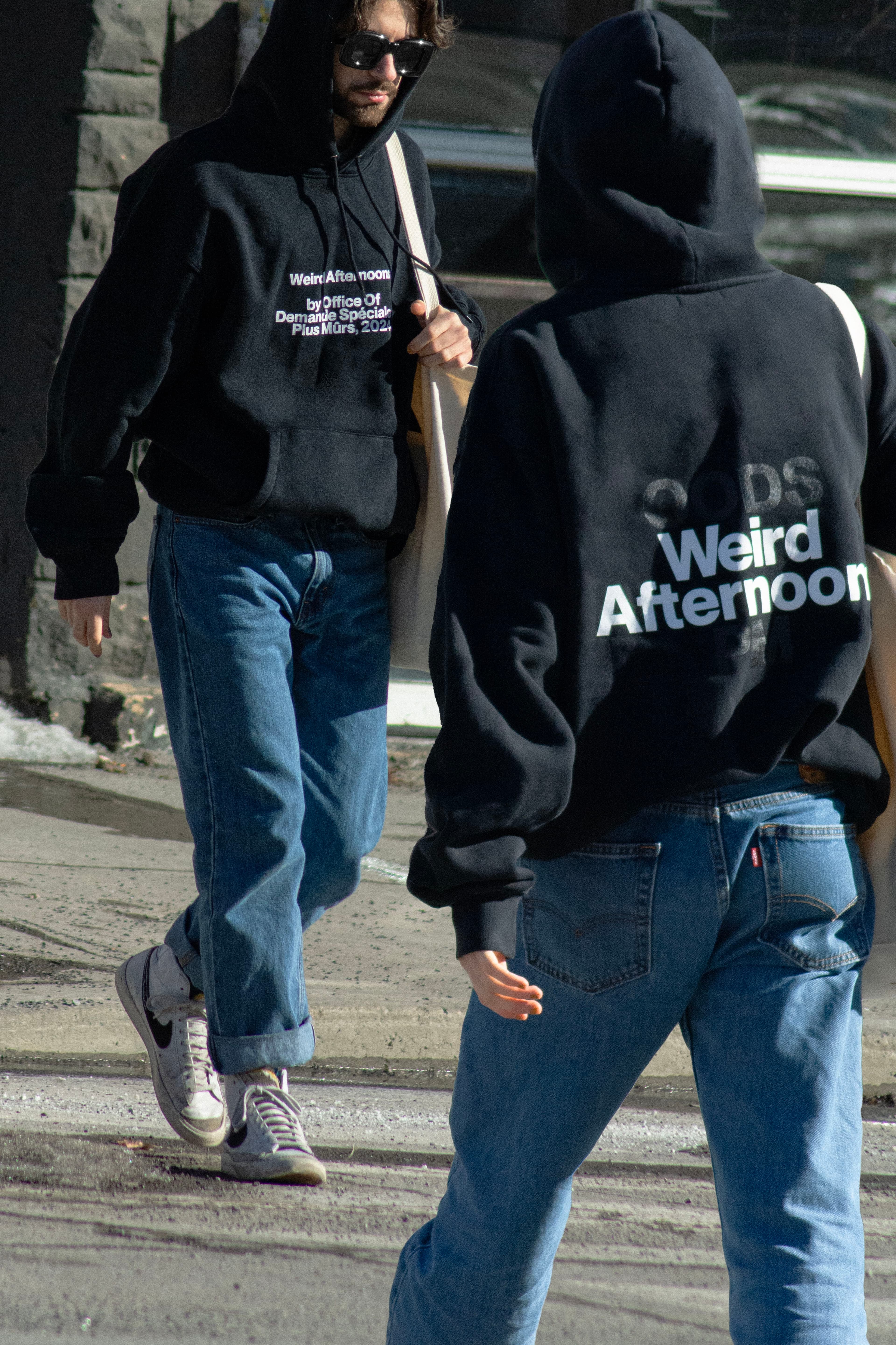 Two people wearing matching black branded hoodies with words "Weird Afternoon" while walking outdoors.
