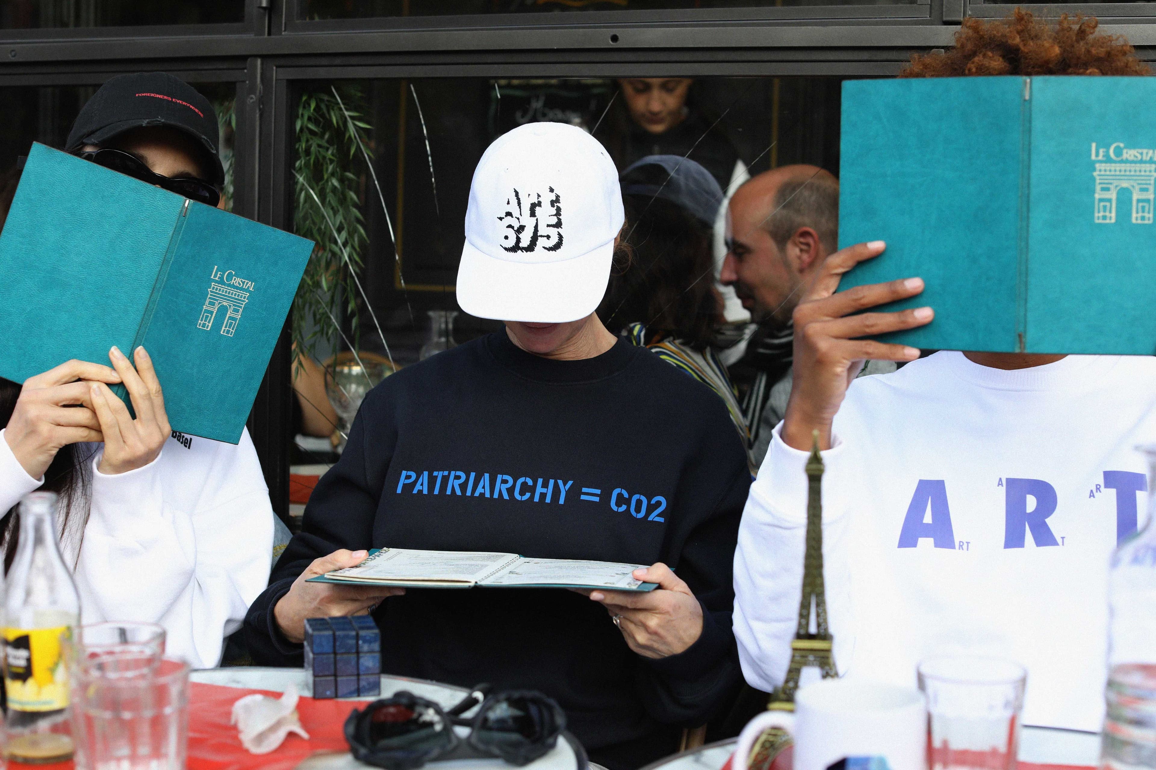 Three people sitting at a table, covering their faces with menus. One wears a hat and shirt with printed text. Drinks and a small Eiffel Tower are on the table.