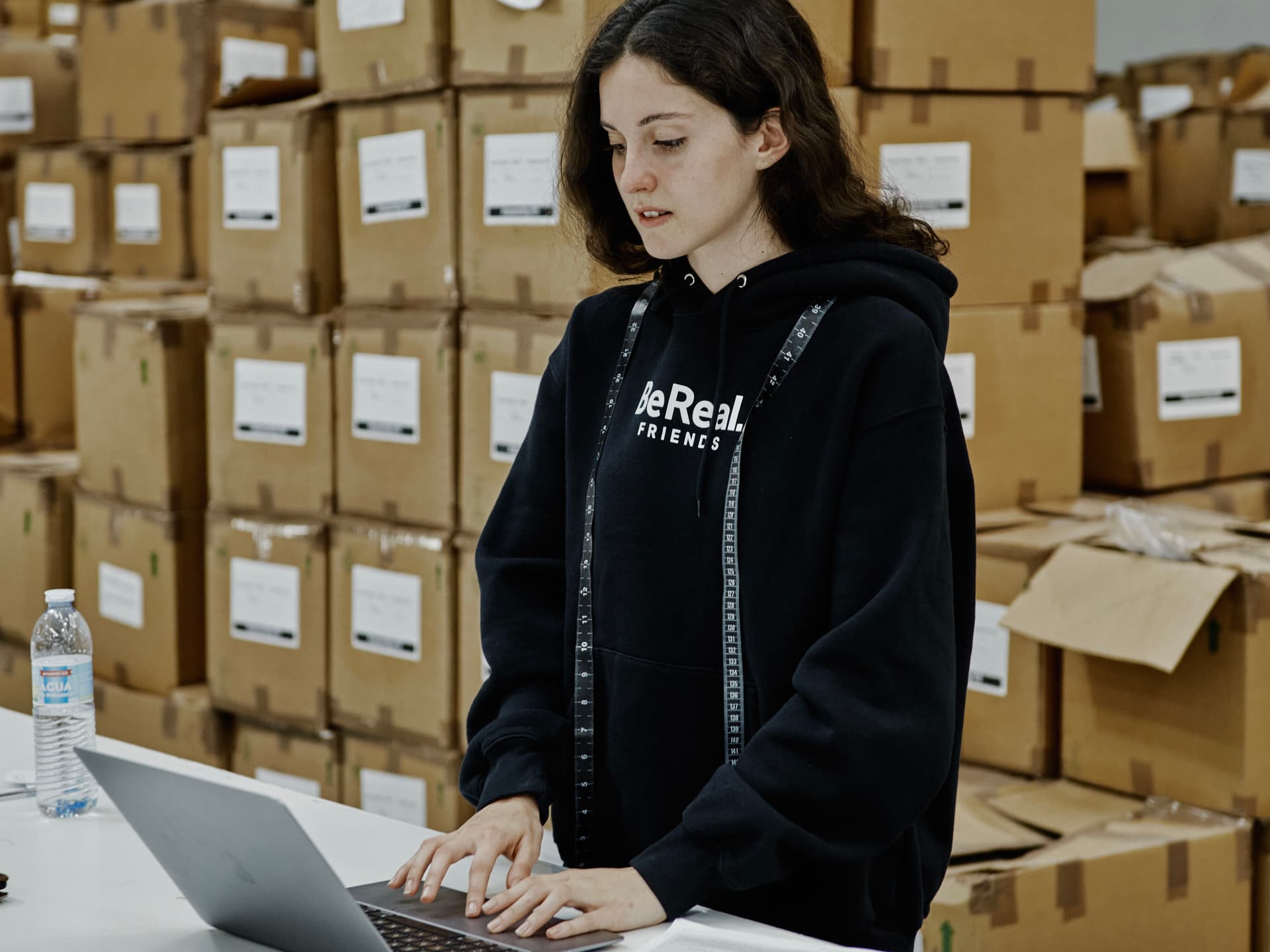 A woman in a black hoodie works on a laptop inside a warehouse, surrounded by stacks of cardboard boxes during the merchandise production process.