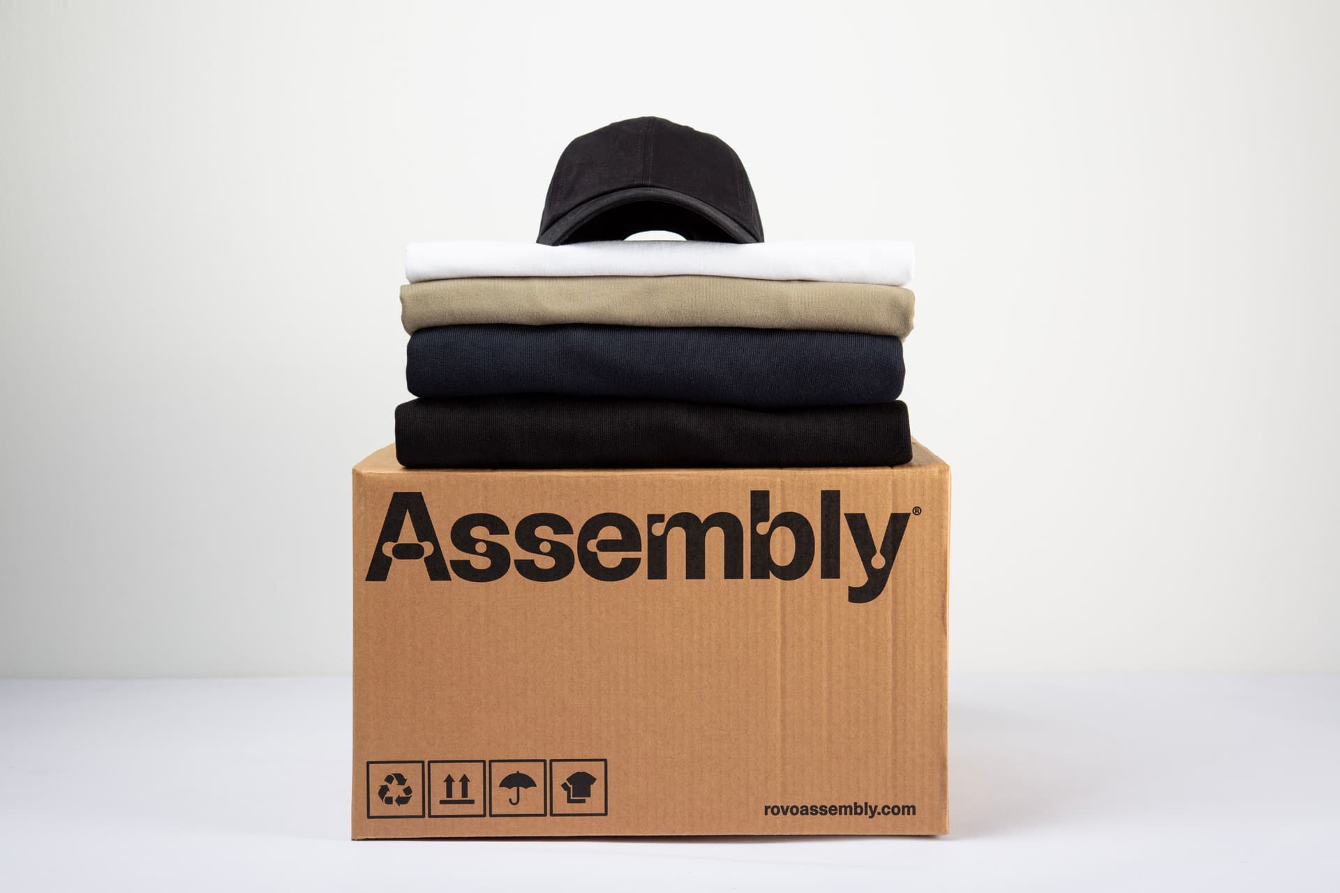 Stack of folded garments and a cap placed on top of an Assembly shipping box, representing delivery and distribution of custom merch.