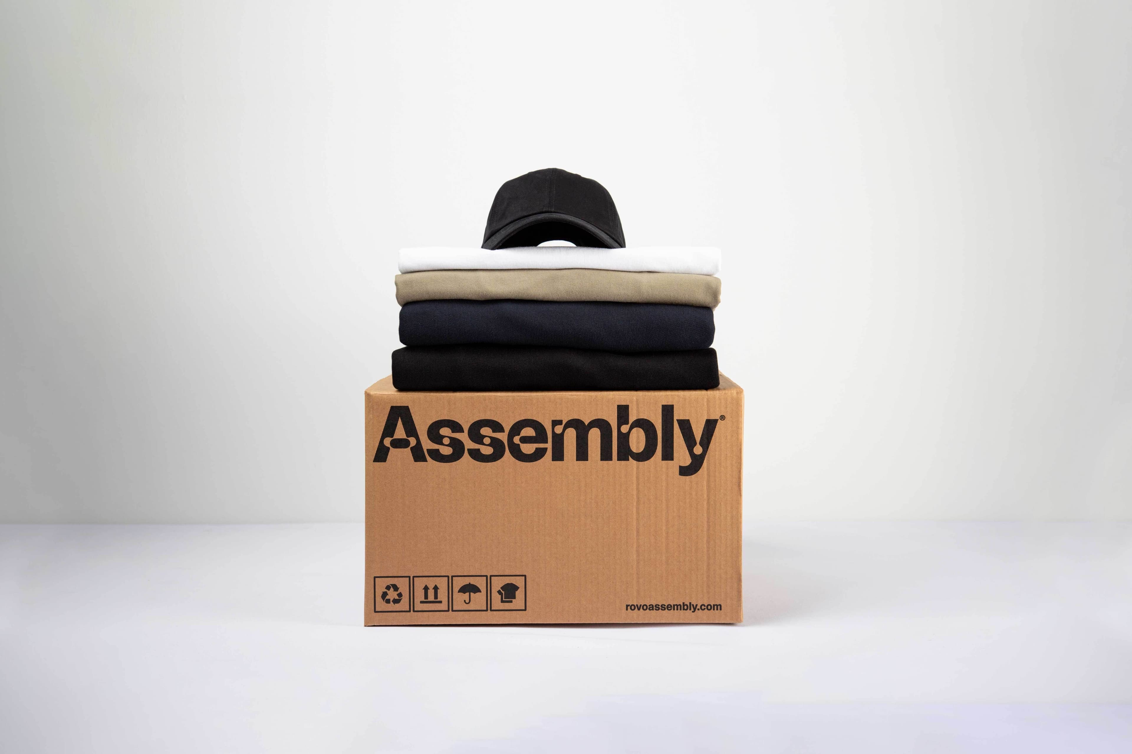 Stack of folded garments and a cap placed on top of an Assembly shipping box, representing delivery and distribution of custom merch.