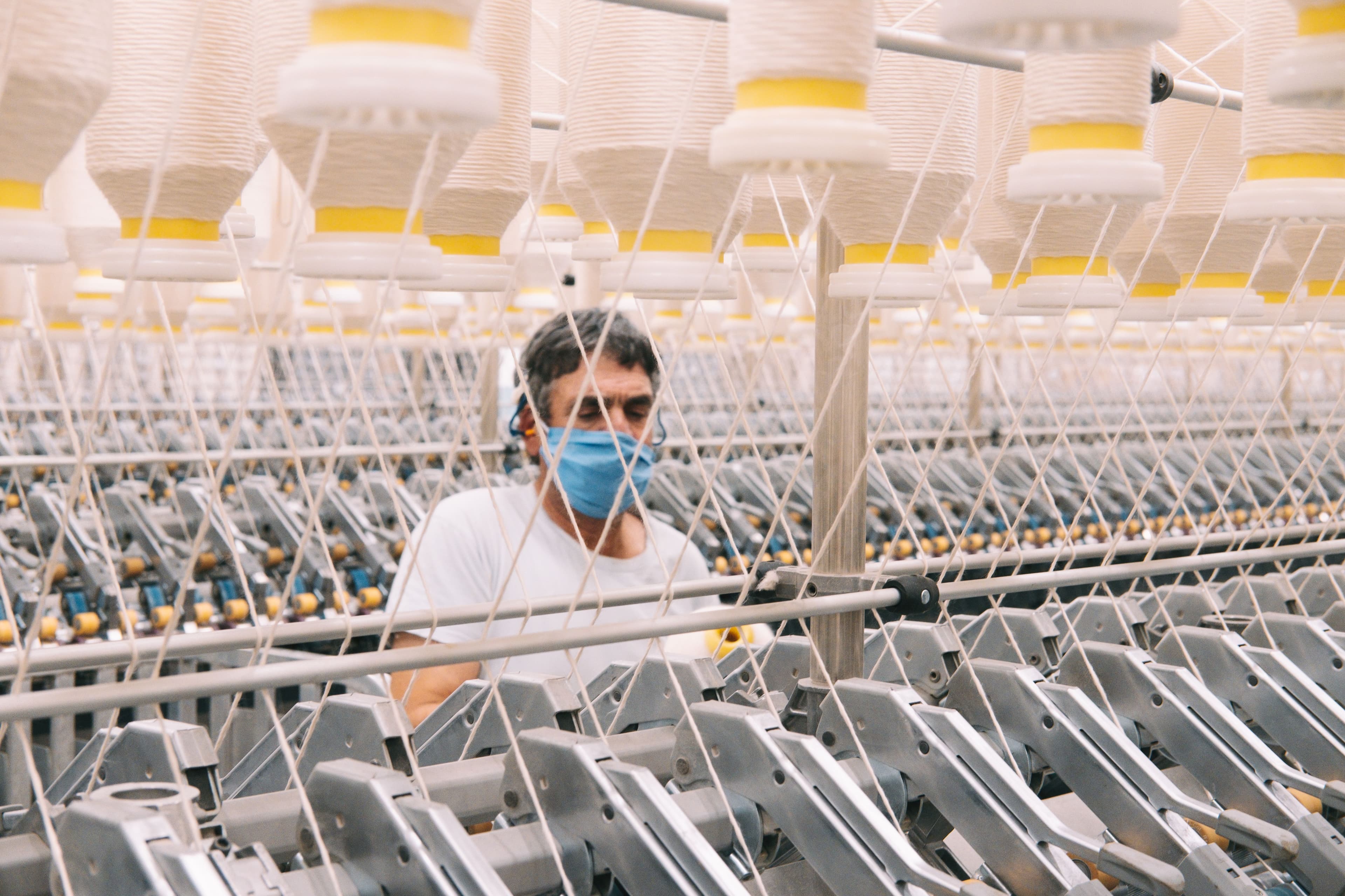 A textile worker wearing a mask operates machinery surrounded by rows of yarn spools in a fabric production facility.