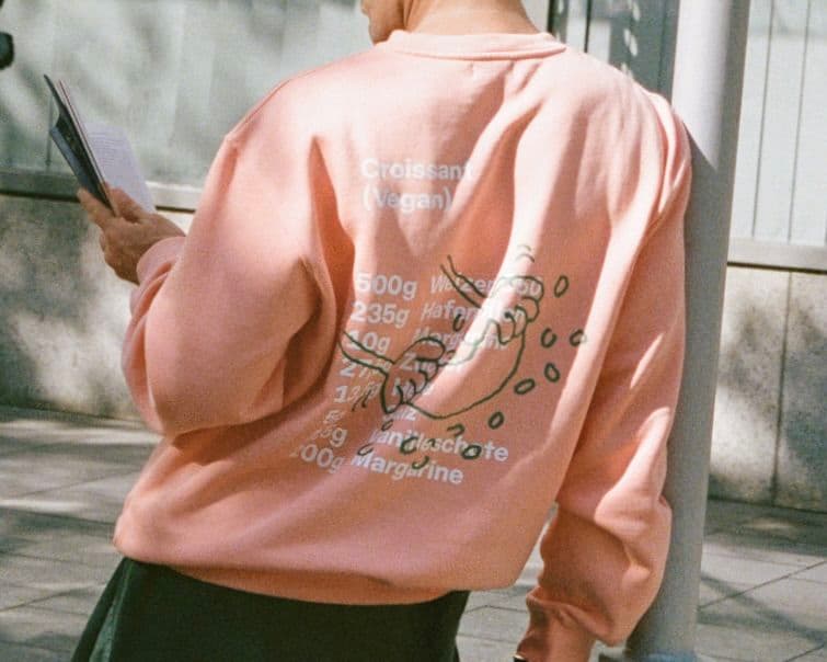 Person in a pink sweatshirt with croissant recipe print on the back, holding a book, standing on a sunlit street.