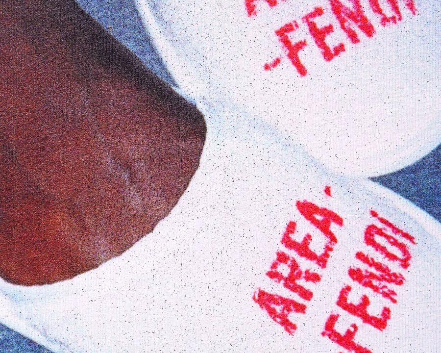 Close-up of white slippers with red text "AREA FENDI" on them, worn by a person with dark skin, against a gray background.