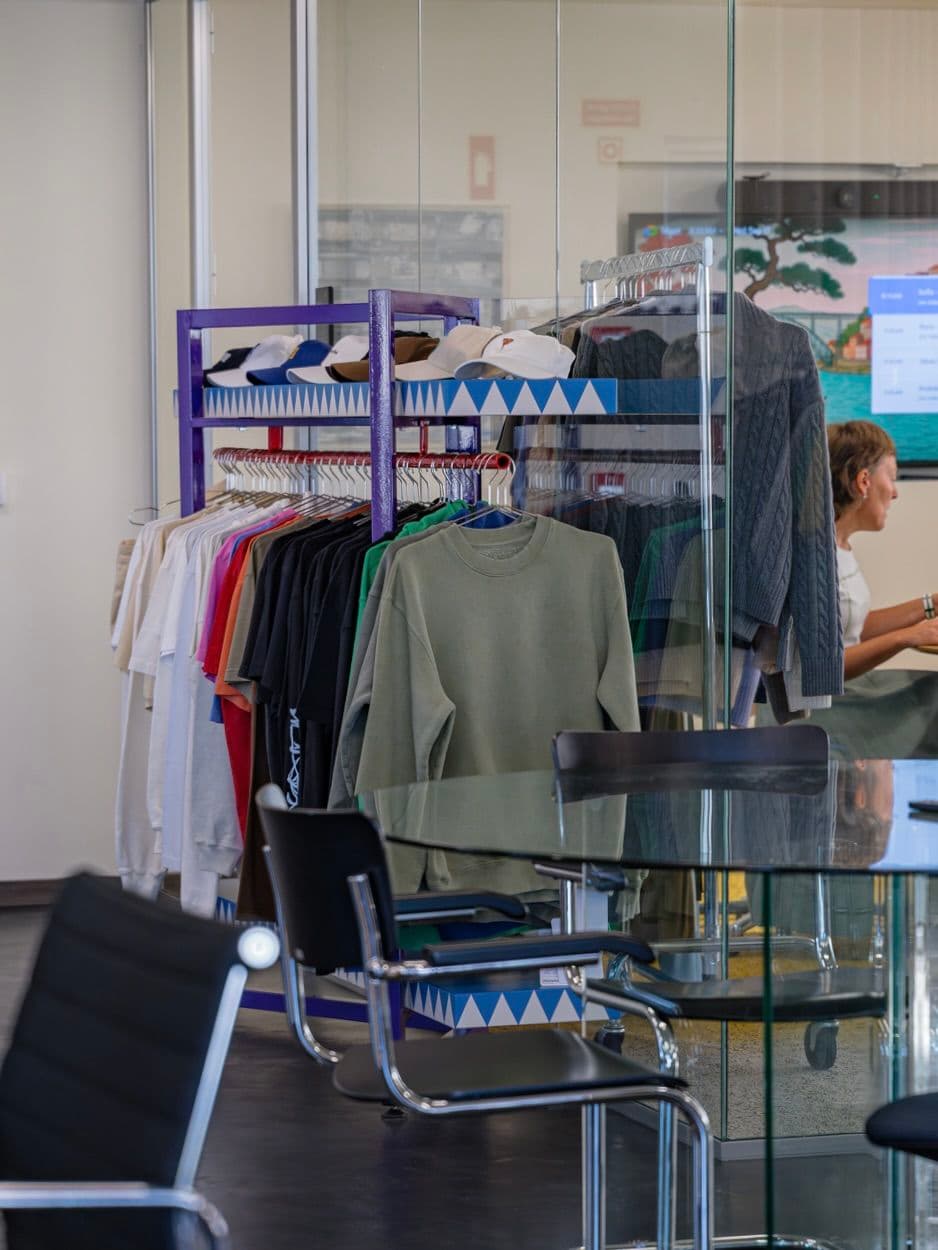 Clothing rack with various shirts, sweatshirts, and caps next to a glass-walled meeting room with black chairs and a person seated inside.